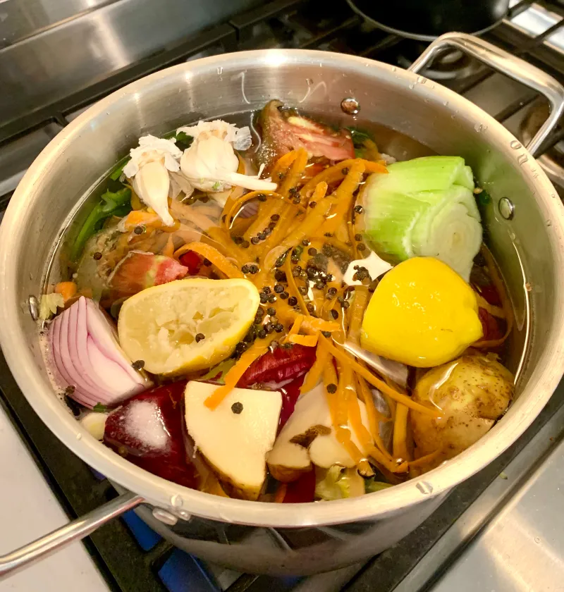 A pot filled with a variety of vegetables and aromatics, including garlic, lemon, carrots, potatoes, onions, and spices, simmering on a stove.